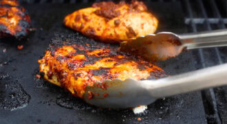 A pair of metal tongs holds a piece of grilled, charred chicken breast on a hot grill, with another piece of grilled chicken cooking beside it.