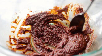 A spoon mixing thick dark brown marinade in a clear glass bowl, with remnants of the mixture visible on the sides.