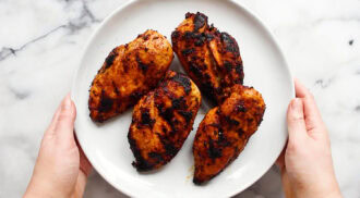 Hands holding a white plate with four pieces of grilled, seasoned chicken breast on a marble surface.