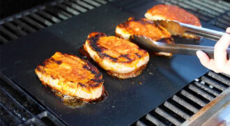 Four pieces of seared pork chops are cooking on a flat grill plate, with a hand using metal tongs to turn one piece.