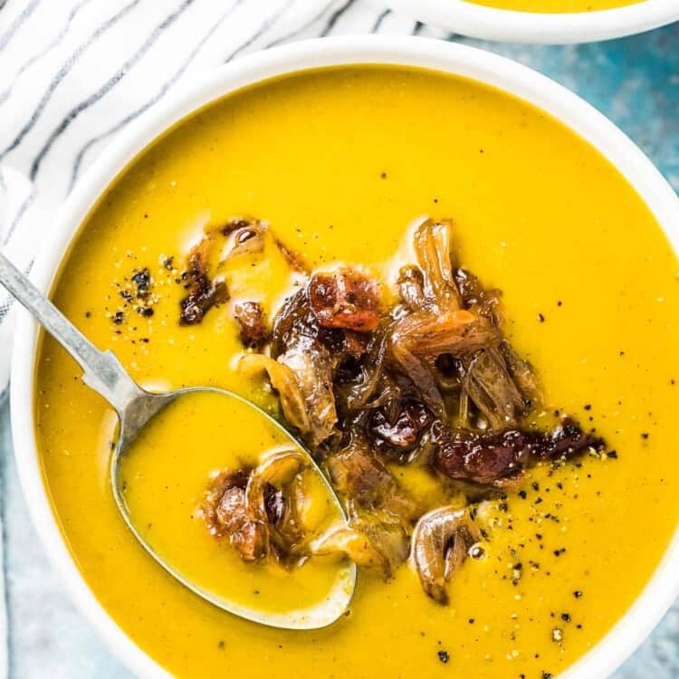 A white bowl with a spoon in it filled with this pumpkin soup and topped with maple bacon onions.