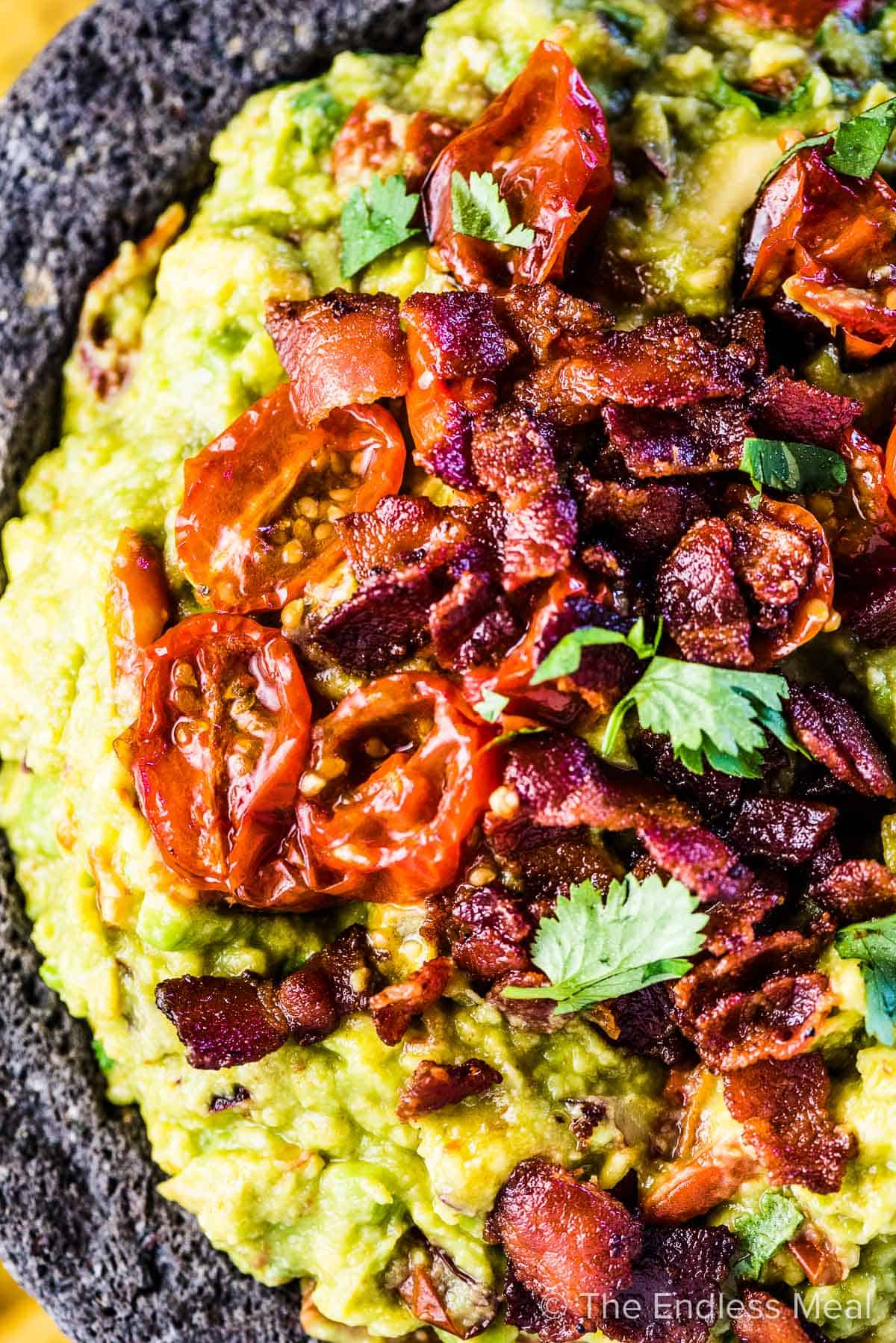 A close up of a serving bowl filled with bacon guacamole and topped with crispy bacon and roasted tomatoes. 