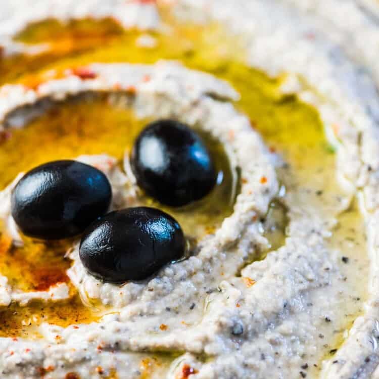 A close up of this white bean dip in a bowl with a few olives on top.