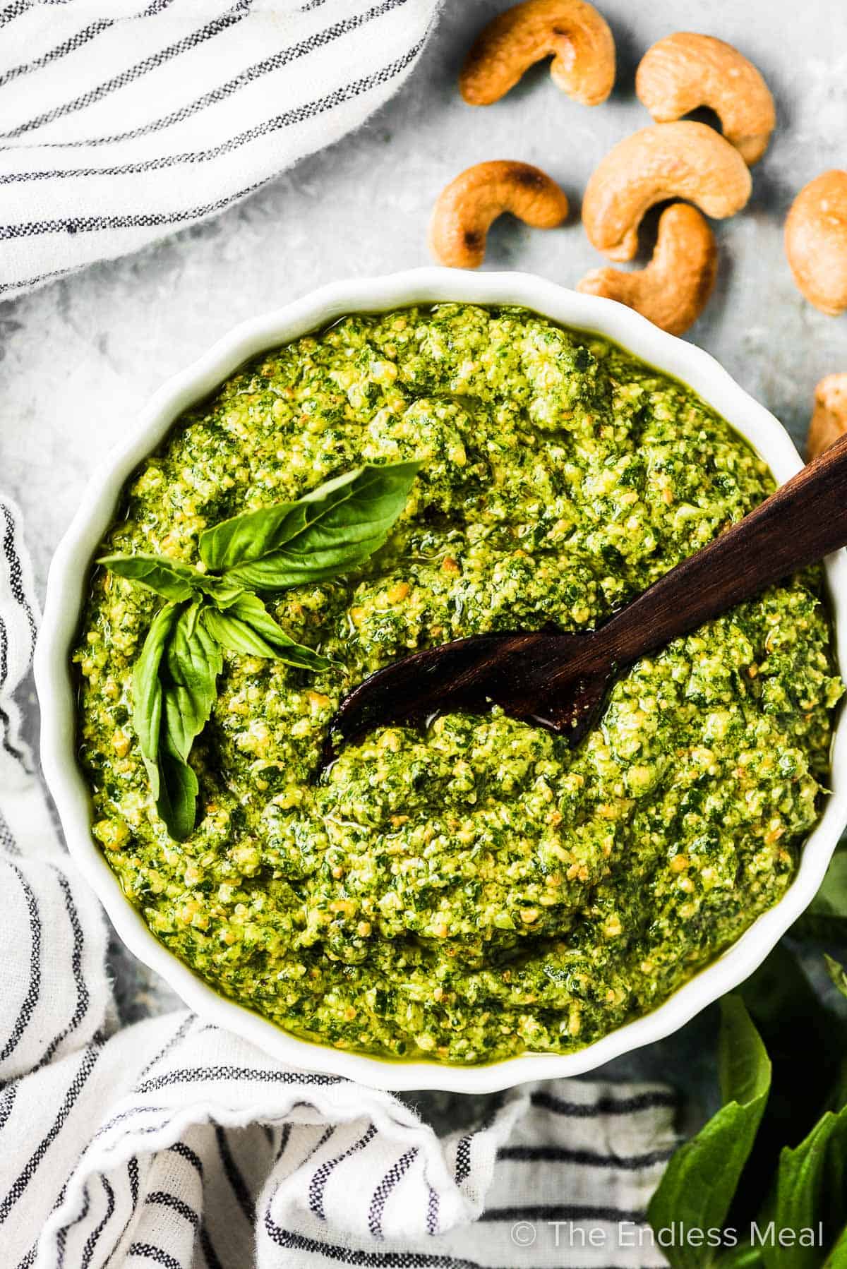 A white bowl of Whole30 pesto with a small wooden spoon in it and some cashews on the side.