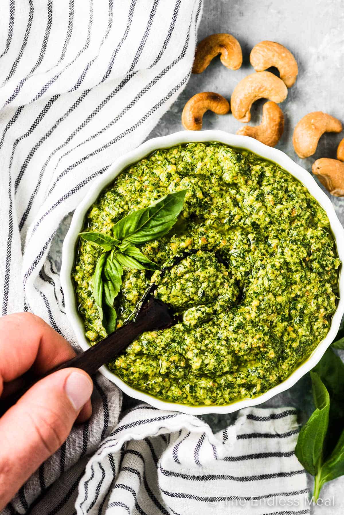 A hand holding a spoon taking a scoop of dairy free pesto out of a white bowl.