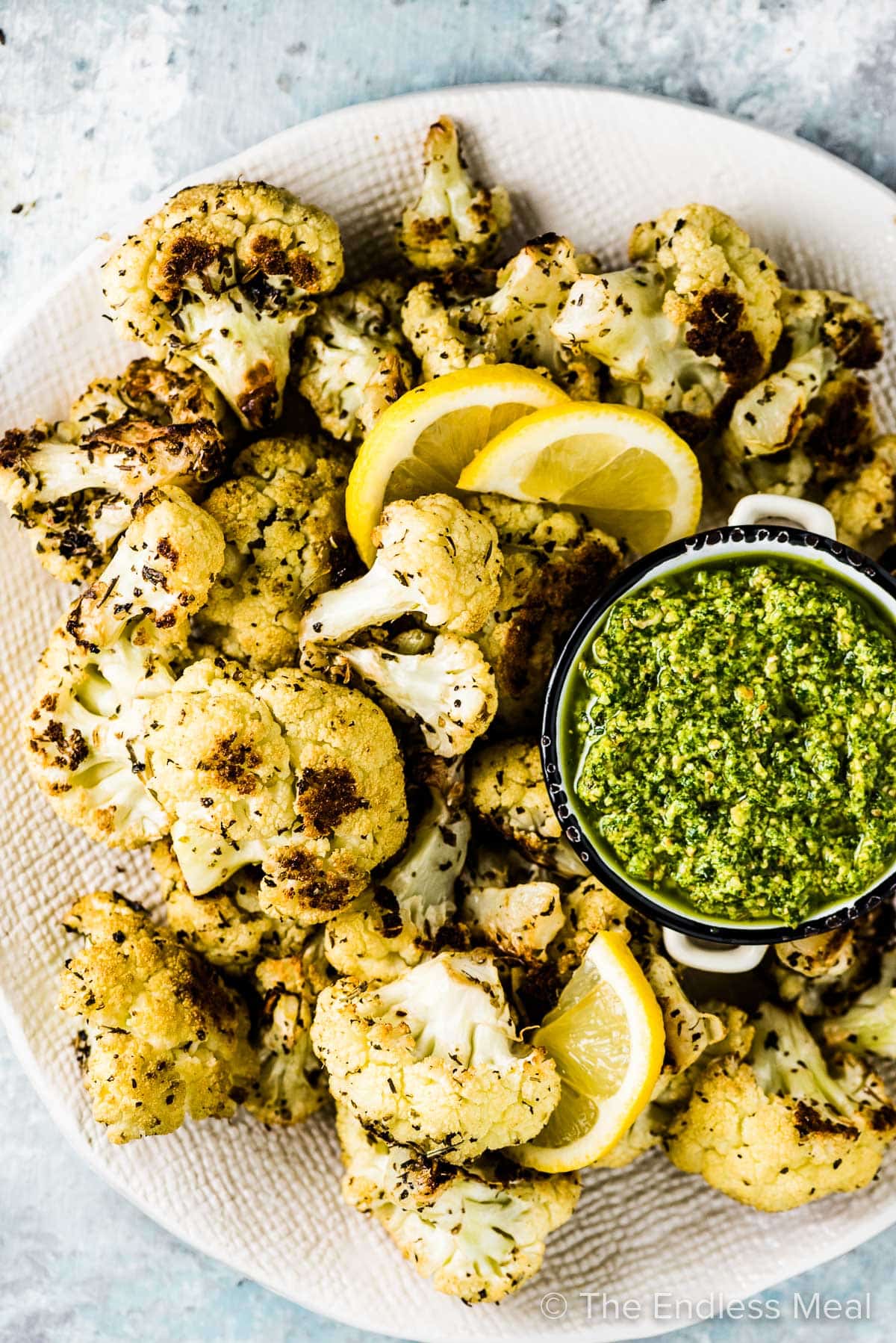 A white serving plate filled with roasted Italian cauliflower with a bowl of pesto and some lemon slices.
