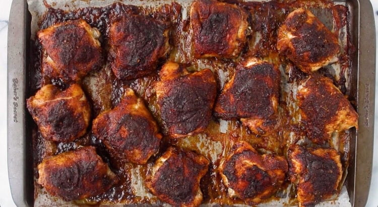 A baking sheet topped with fourteen cooked, seasoned chicken thighs, browned and surrounded by juices on a metal tray.