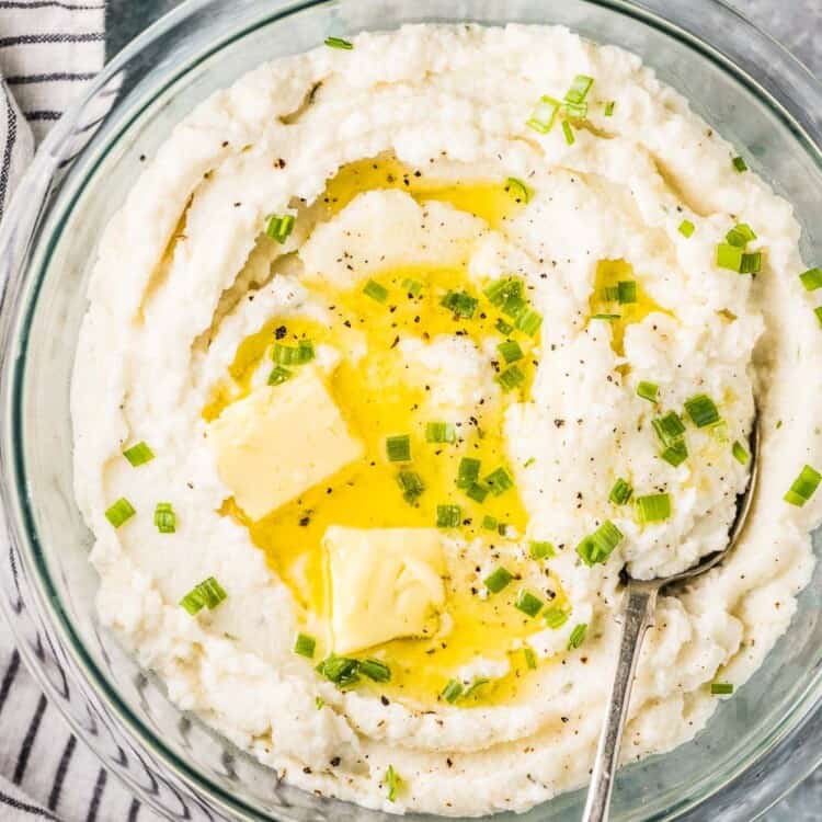 A bowl of the best cauliflower mashed potatoes with butter and chives on top and a spoon in it,