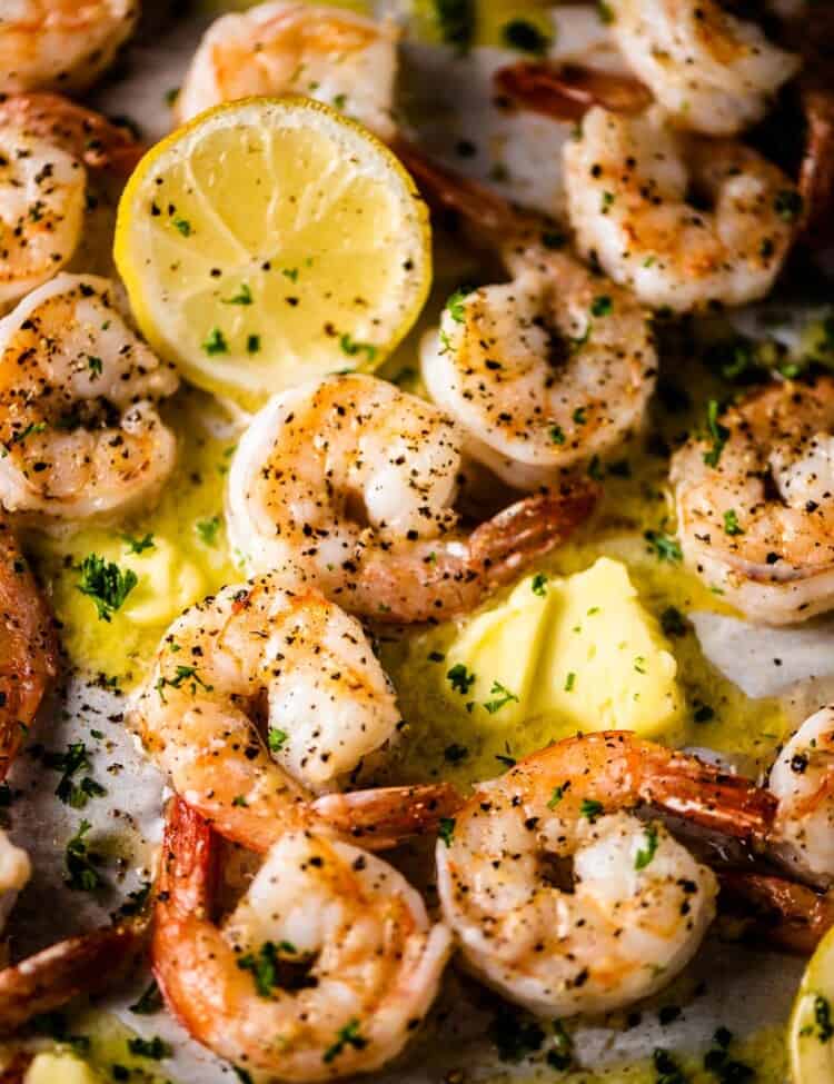Garlic butter baked shrimp on a tray with slices of lemon and pats of butter.