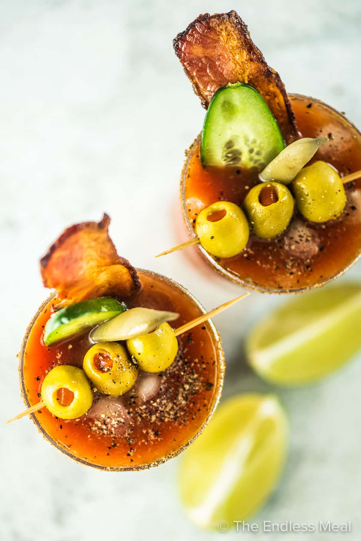 Two Bloody Caesars garnished with bacon, cucumber, pickled beans, and olives.