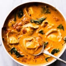 A white bowl of creamy creamy tomato tortellini soup with spinach and a spoon in the bowl.