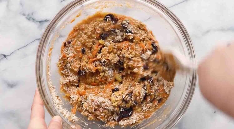 A hand stirs a mixture of oats, raisins, grated carrots, and batter in a glass bowl on a marble countertop—perfect for making wholesome carrot banana muffins.