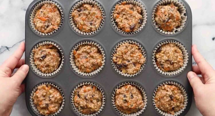 A muffin tin holds twelve paper liners filled with unbaked carrot banana muffins batter, with hands holding each side of the tin on a marble surface.