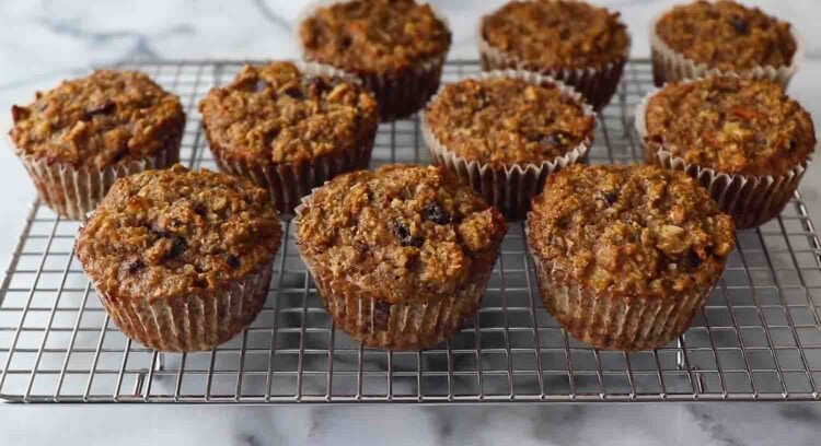 Eleven carrot banana muffins with a brown, textured top are cooling on a wire rack placed on a marble surface.