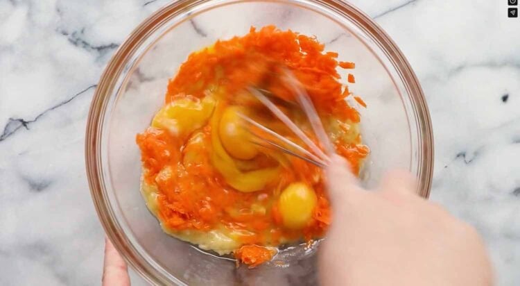 A hand whisks eggs, grated carrots, and mashed banana in a clear glass bowl on a marble surface—fresh ingredients for delicious carrot banana muffins.