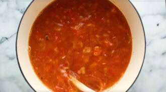 A pot of creamy tortellini soup with a tomato base is being stirred with a white spoon on a white marble countertop.