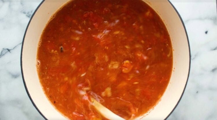 A pot of creamy tortellini soup with a tomato base is being stirred with a white spoon on a white marble countertop.