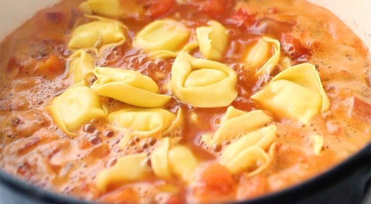 Tortellini pasta is cooking in a tomato-based sauce, with bubbles forming on the surface—perfect for transforming into a creamy tortellini soup.