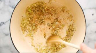 Chopped onions and herbs are being sautéed in a large light-colored pot with a wooden spoon on a marble countertop, the first step in preparing creamy tortellini soup.