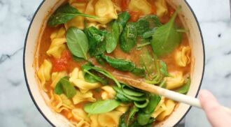 A pot of creamy tortellini soup with tomato broth and fresh spinach is being stirred with a wooden spoon.