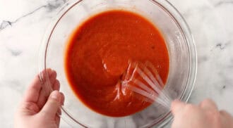 A person whisks a bowl of red sauce for peanut butter chicken in a clear glass mixing bowl on a marble surface.