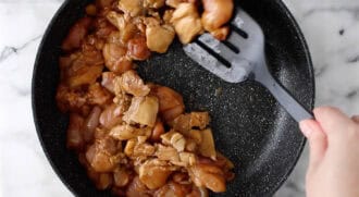 A hand stirs pieces of marinated peanut butter chicken in a black frying pan with a black slotted spatula.
