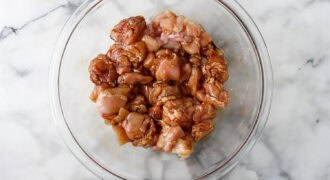 Raw chicken pieces marinated in a rich peanut butter chicken sauce sit in a clear glass bowl on a white marble surface.