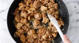 Diced chicken pieces being stir-fried in a black skillet with a white spatula on a marble surface, ready to be transformed into delicious peanut butter chicken.