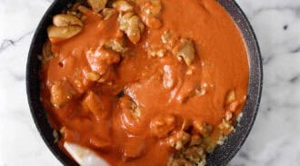 Chunks of chicken cooking in a frying pan covered with a thick orange-red tomato-based sauce, perfect for making peanut butter chicken.
