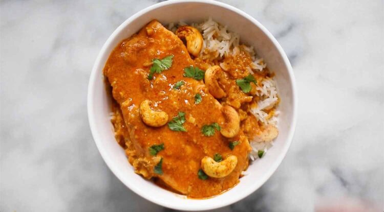 A white bowl filled with rice, chicken in orange curry sauce, and garnished with cashews and fresh cilantro on a marble surface, reminiscent of a vibrant salmon curry presentation.