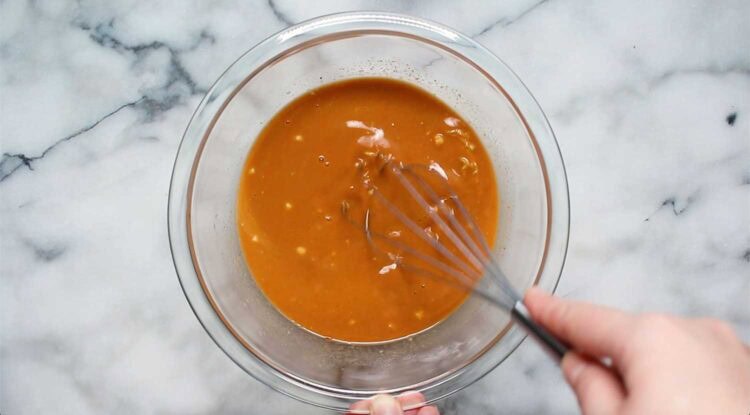 A hand uses a whisk to mix a brown salmon curry sauce with visible chunks in a glass bowl on a marble countertop.