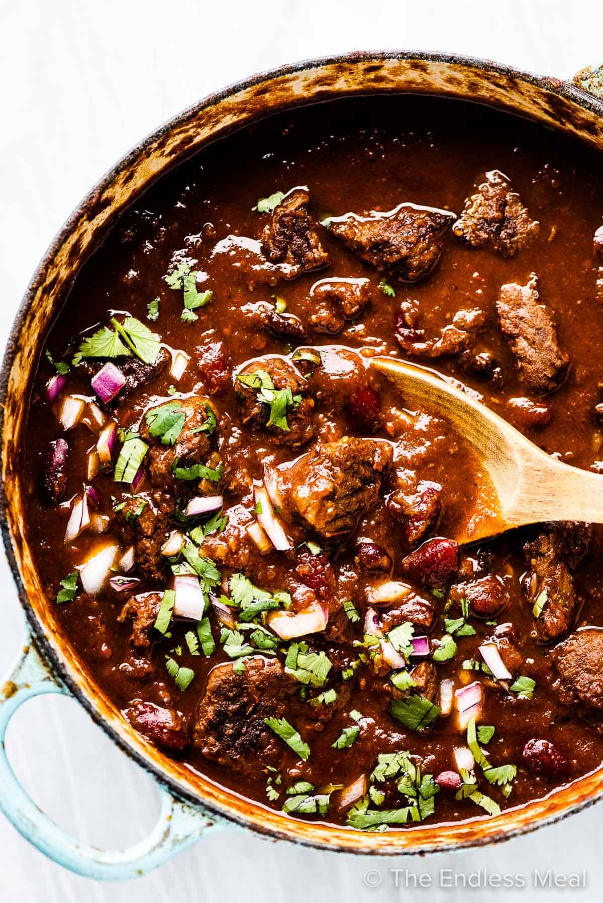 A spoon taking a scoop of the best steak chili out of a pot.