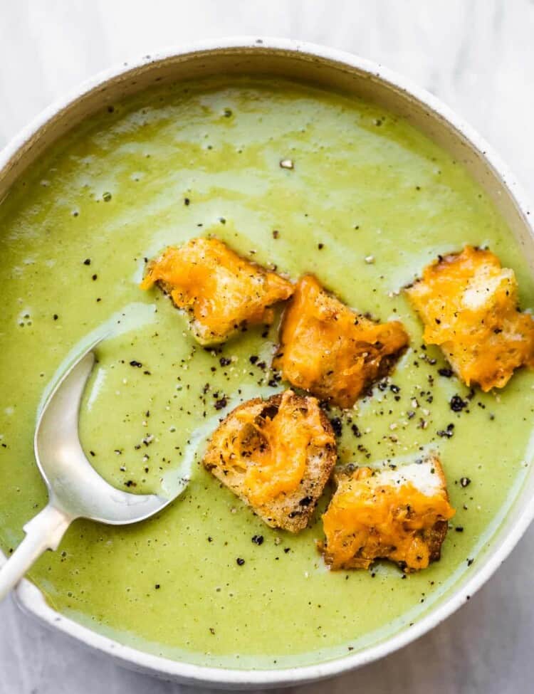 A spoon taking a scoop of asparagus soup from a white bowl topped with croutons.