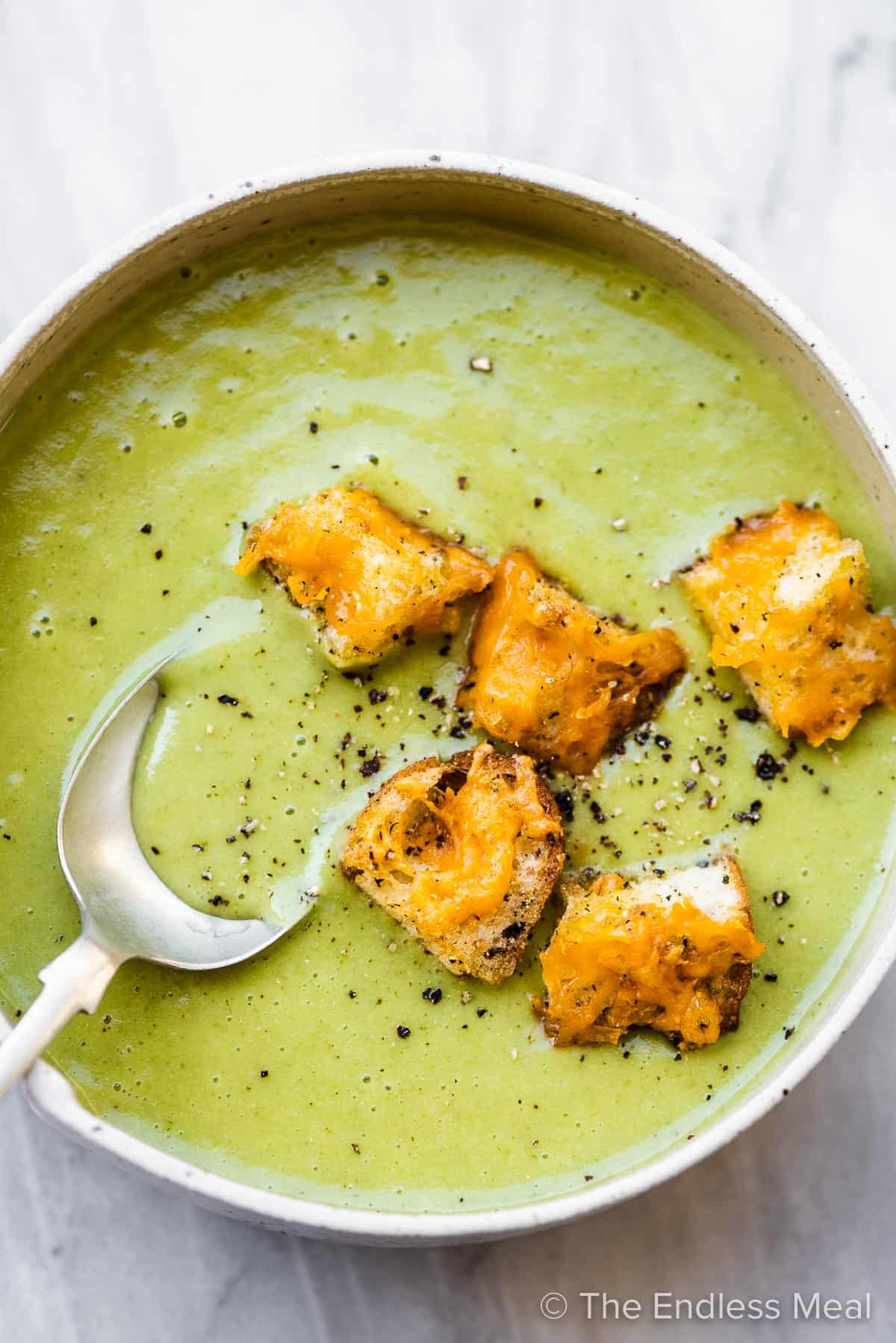 A spoon taking a scoop of asparagus soup from a white bowl topped with croutons.