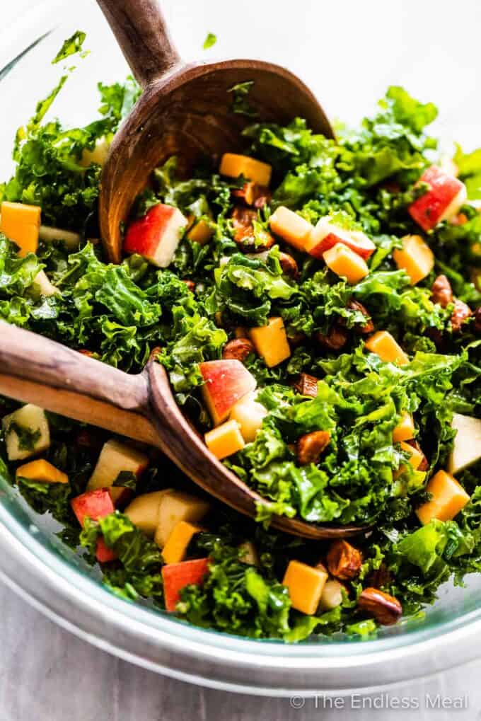 A close up of kale apple salad in a glass bowl with wooden salad tongs.