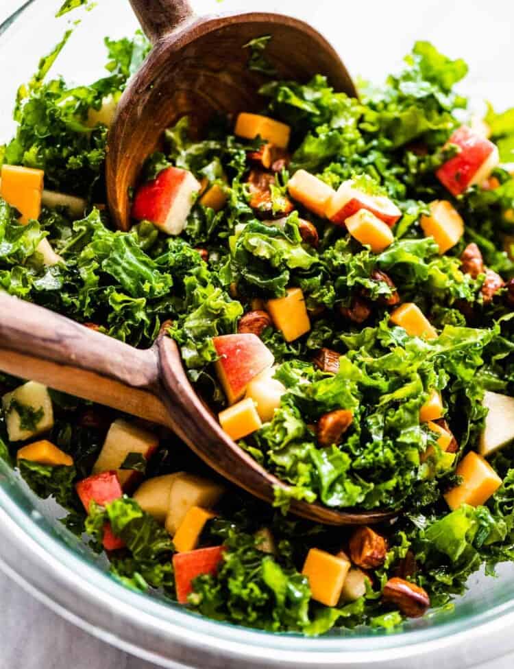 A close up of kale apple salad in a glass bowl with wooden salad tongs.