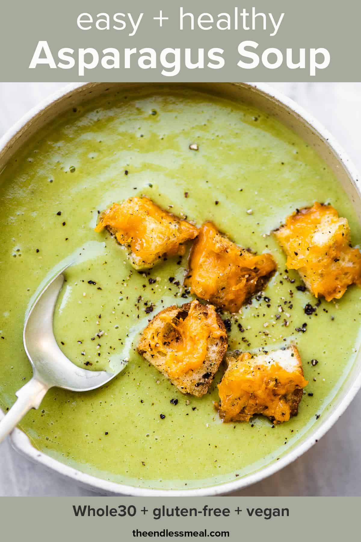 A spoon taking a scoop of asparagus soup from a white bowl topped with croutons and the recipe title on top of the picture.