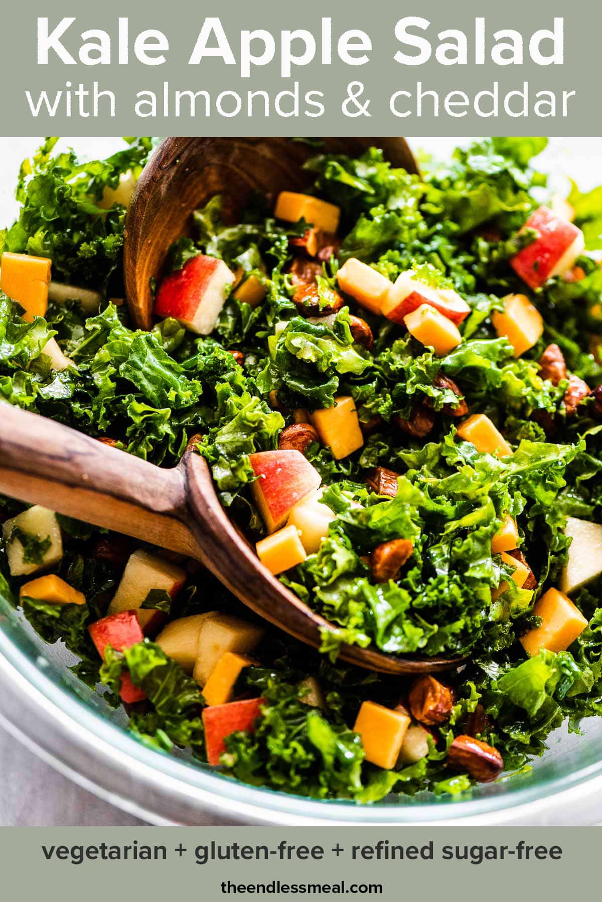 A close up of kale apple salad in a glass bowl with wooden salad tongs and the recipe title on top of the picture.