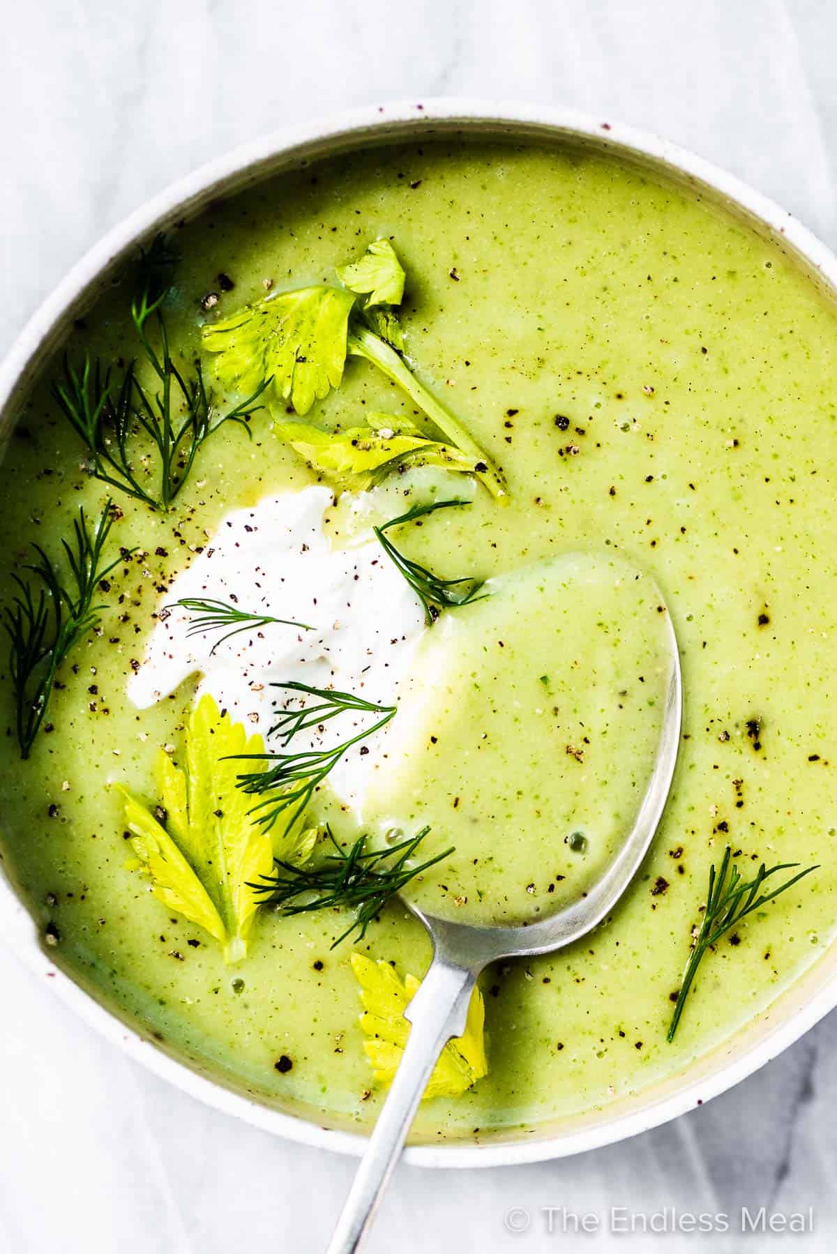 Celery soup in a white bowl with a spoon and some sour cream and dill on top.