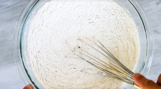 A hand holds a whisk mixing flour in a large glass bowl, preparing dough for no yeast bread.