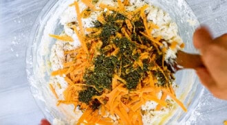 A glass bowl containing a mix of shredded cheese, herbs, and flour for a no yeast bread is being stirred with a wooden spoon by hand.