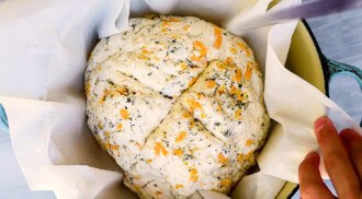 A round loaf of unbaked no yeast bread with herbs and cheese sits in a parchment-lined baking dish, with a hand scoring the top.