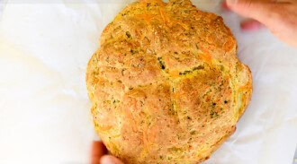 A round loaf of golden-brown no yeast bread with a textured crust rests on white parchment paper; hands are positioned near the bread.