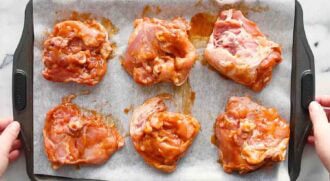 Six uncooked, marinated baked BBQ chicken pieces are arranged on a parchment-lined baking tray, ready for cooking.