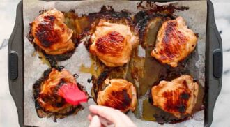 A baking sheet lined with parchment paper holds six baked BBQ chicken thighs, with a hand brushing sauce onto one of the pieces.