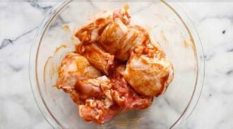 Raw chicken pieces coated in a reddish baked BBQ chicken marinade sit in a clear glass bowl on a white marble surface.