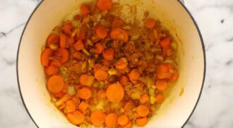 Chopped carrots, onions, and spices simmer for a curried cauliflower soup in a white pot on a marble surface.