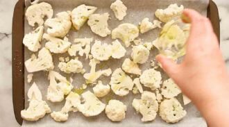 A hand drizzles oil over raw cauliflower florets spread on a parchment-lined baking sheet, the perfect start for a flavorful curried cauliflower soup.