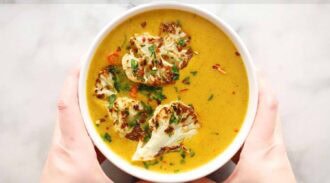 A pair of hands holds a bowl of creamy curried cauliflower soup, topped with roasted cauliflower florets and sprinkled with chopped herbs and red pepper flakes.