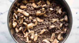 A close-up of cooked ground beef stir fry with sliced mushrooms in a metal pan on a marble surface.